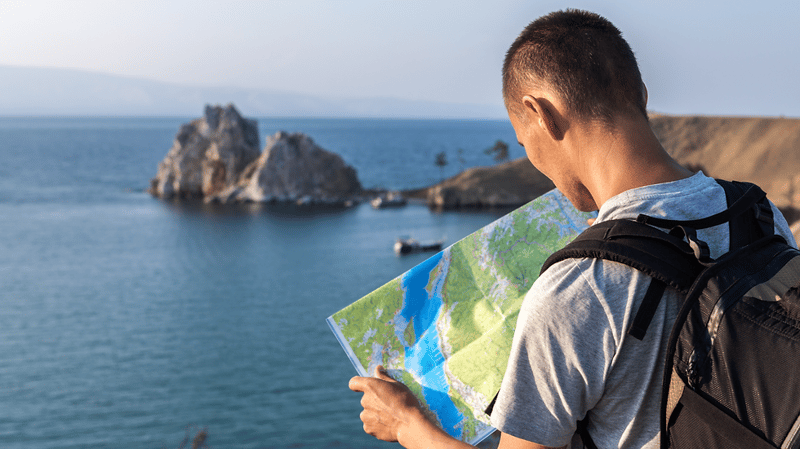 A man holding a map overlooking water in a bay. Fog covered peaks are visible on the opposite side.